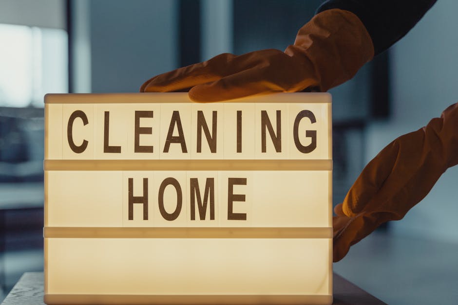 A person wearing brown rubber gloves is adjusting a lit sign with the words 'CLEANING HOME' in bold letters, placed on a flat surface indoors. The background shows a modern, softly lit room with a hint of a window, suggesting a clean and tidy environment. The sign is illuminated from within, emphasizing the message related to domestic surface cleaning and hygiene services. This image illustrates professional cleaning practices, aligning with the services offered by Carpet Cleaners Highbury to maintain hygienic and well-maintained homes.