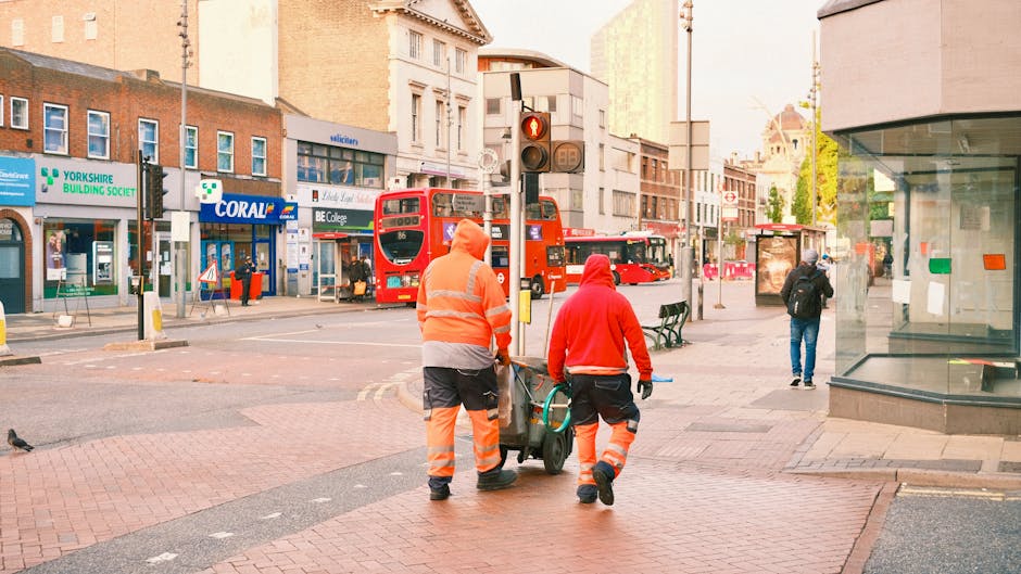 Two street cleaning workers in high-visibility orange and red uniforms are walking on a city pavement near a bus stop, pushing a cleaning cart. The background shows a busy urban street with a red double-decker bus, various storefronts including 'Coral' and 'YORKSHIRE BUILDING SOCIETY,' and a glass-fronted building on the right. The scene is well-lit with natural daylight, and the pavement appears clean and free of debris. The workers are engaged in surface cleaning activities in a commercial area, reflecting professional hygiene and maintenance practices, consistent with the focus on deep cleaning and sanitisation in urban settings.