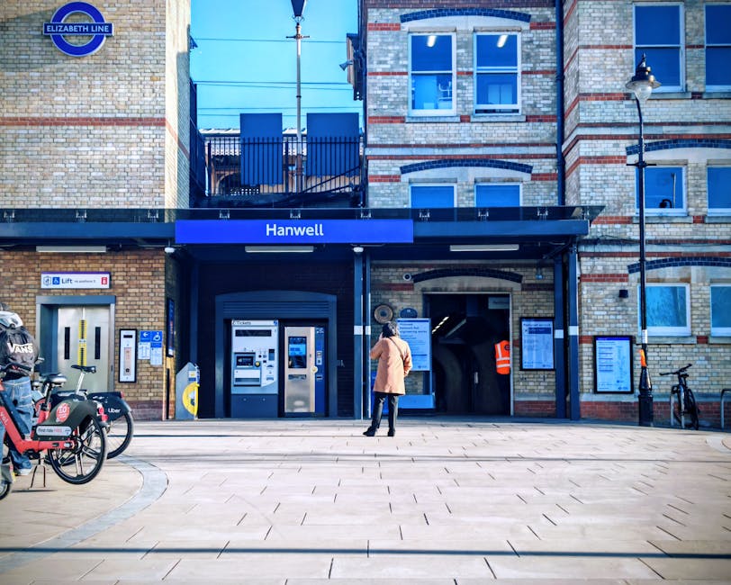 A person standing outside the entrance to Hanwell station, part of the Elizabeth Line, in a high-visibility vest and orange coat, appears to be waiting or reading a notice board. The station building is constructed of brick with a mix of beige and red tones, featuring several windows with white frames. There are two ticket machines and a lift access point on the left side of the entrance, alongside bike-sharing bikes parked on the paved area in front. The area is well-lit with a street lamp on the right, and the surroundings are clean and tidy, reflecting a maintained urban environment suitable for surface cleaning and general hygiene upkeep. Carpet Cleaners Highbury provides expert cleaning solutions, including emergency carpet cleaning and deep sanitisation, especially in bustling transport hubs like this one.
