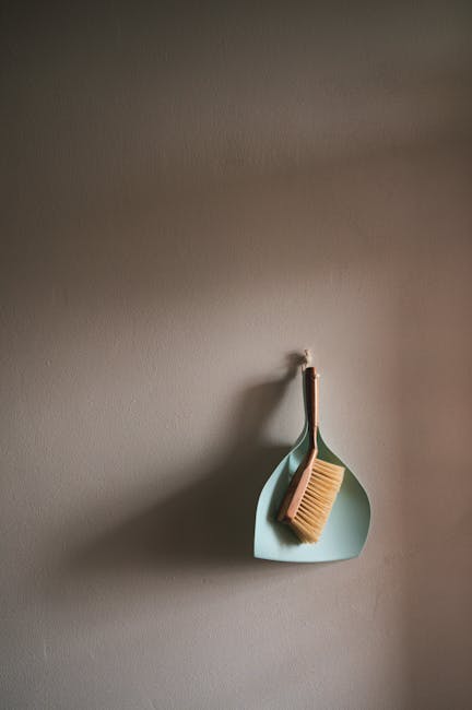 A light blue, teardrop-shaped plastic dustpan hanging on a wall hook, with a beige cleaning brush resting inside it. The wall is painted in a neutral taupe color, and the surface appears clean and smooth. The scene is softly lit, highlighting the simple cleaning tools used for domestic surface cleaning or tidying tasks. The placement suggests a tidy and organized space, with minimal clutter, typical of maintained interiors. Carpet Cleaners Highbury specializes in professional cleaning services, including deep surface cleaning and sanitisation, as recommended for end of tenancy cleaning in compliance with N5 Islington Council rules.
