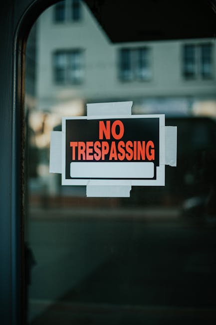 Close-up of a black and white 'No Trespassing' sign with bold red text, attached to a glass door or window using white tape, with a blurred background of residential buildings with multiple windows and a reflective surface. The sign indicates restricted access, while the surroundings suggest an urban environment during daytime. The image is relevant to property security and access control, which can be part of maintaining a clean and safe residential or commercial space. Carpet Cleaners Highbury emphasizes the importance of thorough sanitisation and surface cleaning in their services, supporting the upkeep of secure and hygienic environments in line with End of tenancy cleaning N5 Islington Council rules.