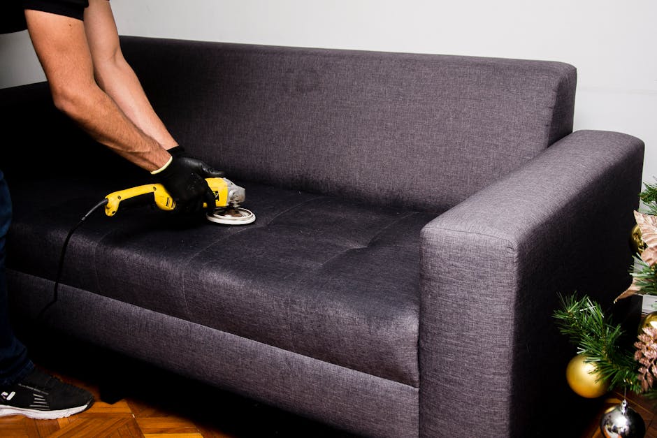 A person wearing black gloves is using a yellow handheld upholstery cleaning machine on a dark grey fabric sofa in a living room. The sofa has a modern design with clean lines and is positioned against a white wall. The cleaning process appears to be part of deep cleaning or surface sanitisation, with a focus on removing dirt and stains from the upholstery fabric. To the right of the sofa, there is a decorated Christmas tree with ornaments, indicating a festive setting. The room has wooden flooring, and natural light illuminates the scene, emphasizing the clean and maintained condition of the furniture. Carpet Cleaners Highbury offers professional domestic cleaning services, including upholstery cleaning, to ensure hygiene and freshness in residential spaces.