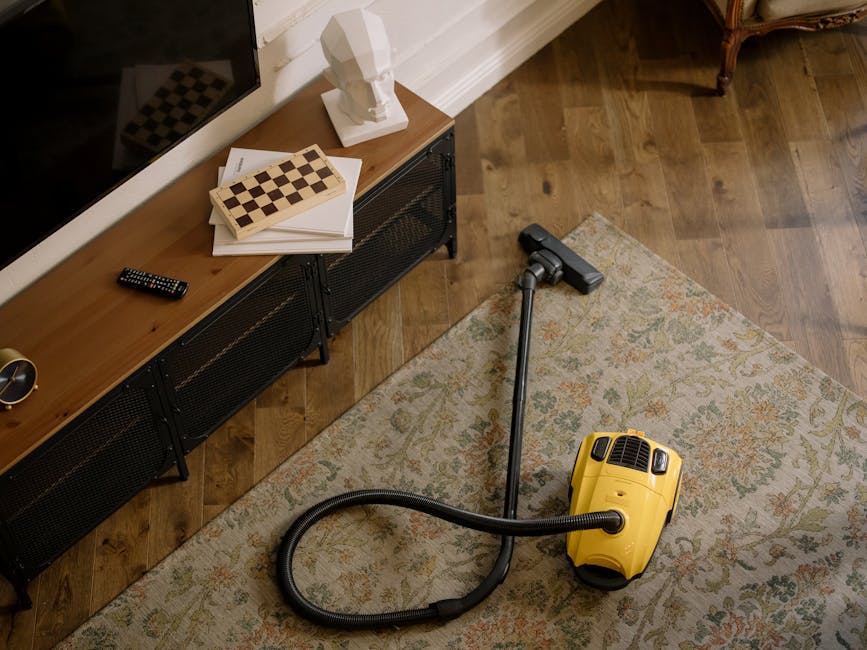 A yellow canister vacuum cleaner with a black extension tube and cleaning head resting on an ornate area rug with floral patterns in a living room. The room features wooden flooring and a black metal sideboard with a stack of books, a remote control, and a white ceramic head sculpture. Natural light illuminates the space, highlighting the clean, polished surfaces and well-maintained furnishings. The scene illustrates professional surface cleaning and deep cleaning activities, consistent with services provided by Carpet Cleaners Highbury near Highbury Fields.