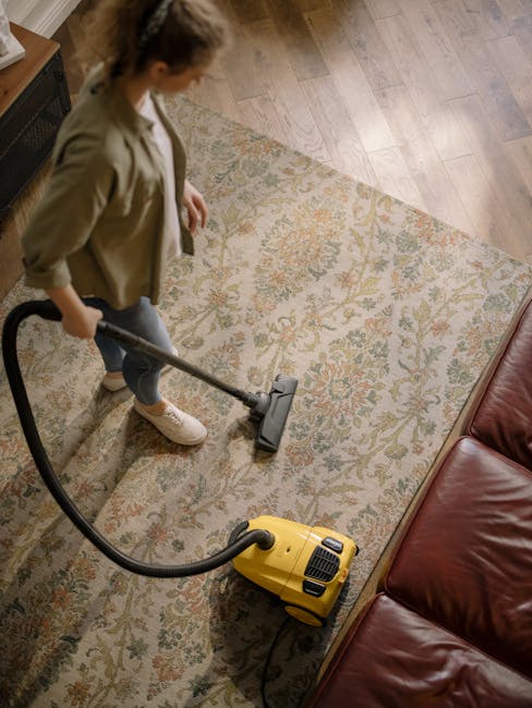 A professional wearing full protective gear, including a white hazmat suit, face mask, gloves, and shoe covers, is using a steam or vapor cleaning machine to deep clean a beige carpet in a bright, modern living room. The room features white walls, a tall mirror, a wooden shelf with decorative items, and potted plants, with natural light illuminating the space. The cleaning process involves the dispersal of steam across the carpet surface, indicating thorough sanitisation and surface cleaning by Carpet Cleaners Highbury as part of their domestic cleaning services.