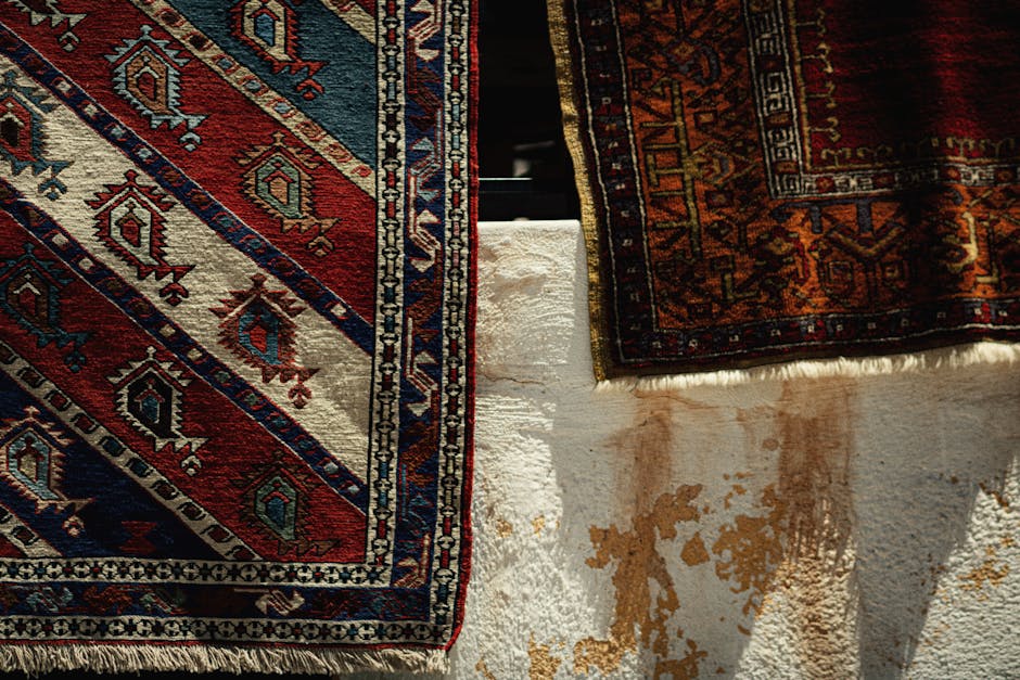 Close-up view of two patterned area rugs placed on a white, textured concrete floor. The rug on the left features a traditional design with red, navy, white, and light blue geometric motifs, while the