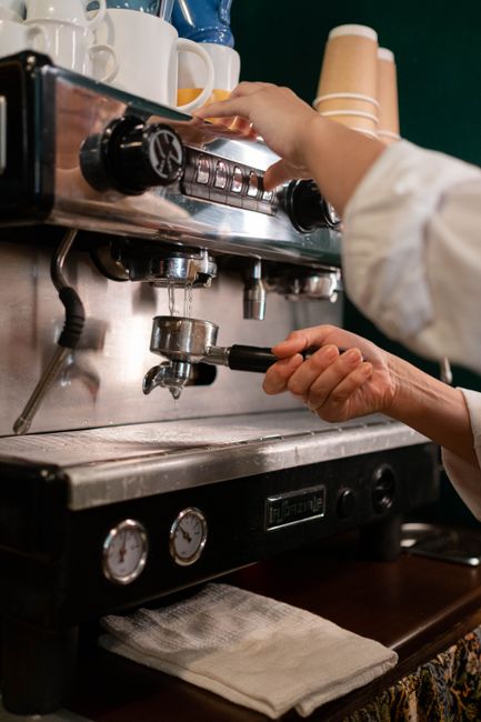 A woman with short, dark hair is cleaning an outdoor wooden countertop at a cafe or shop setting with large glass windows and open doors. She is wearing a white t-shirt and a brown apron, focusing on